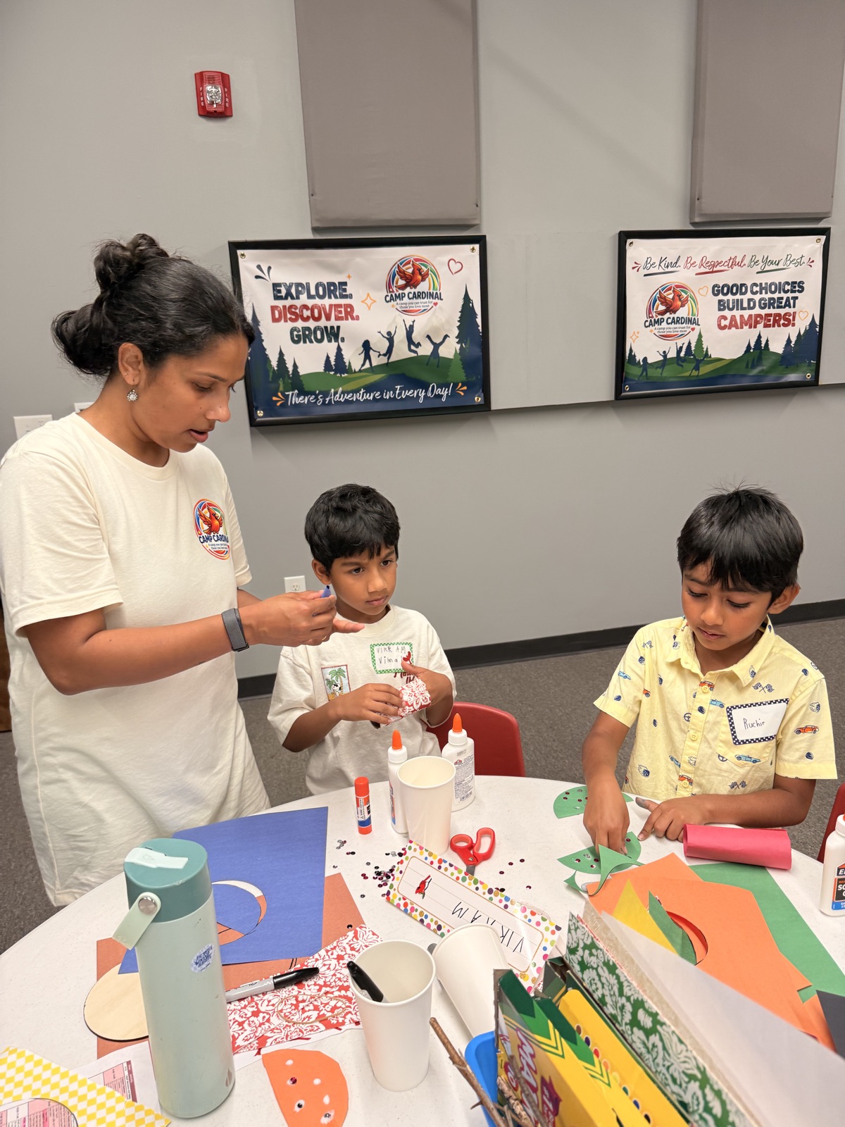 A counselor helps two campers with paper crafts at an activity table